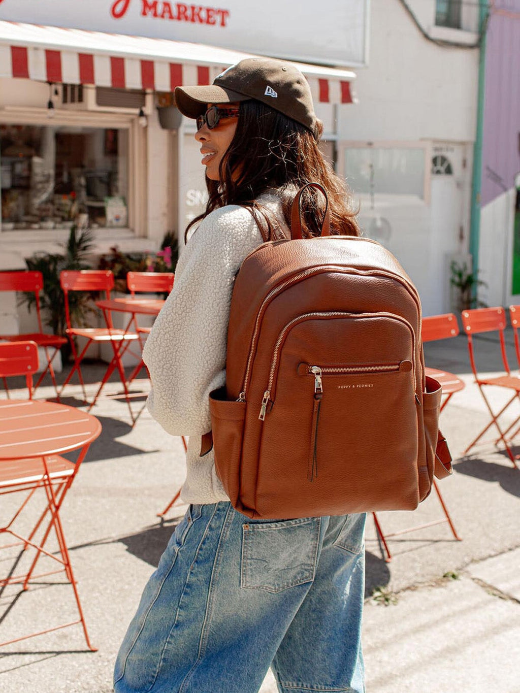 Person wearing a brown backpack in an outdoor setting with red chairs and tables.