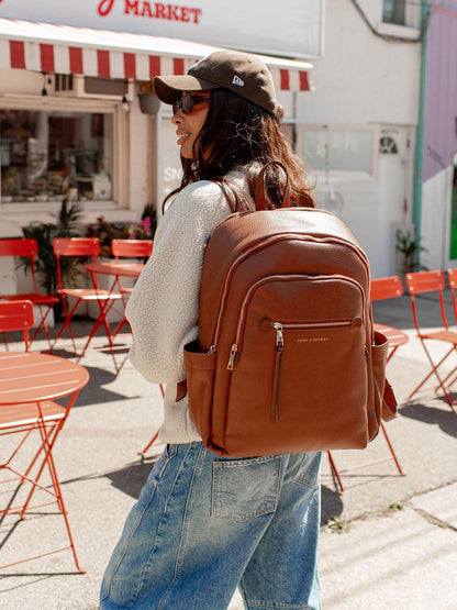 Person wearing a brown backpack in an outdoor setting with red chairs and tables.
