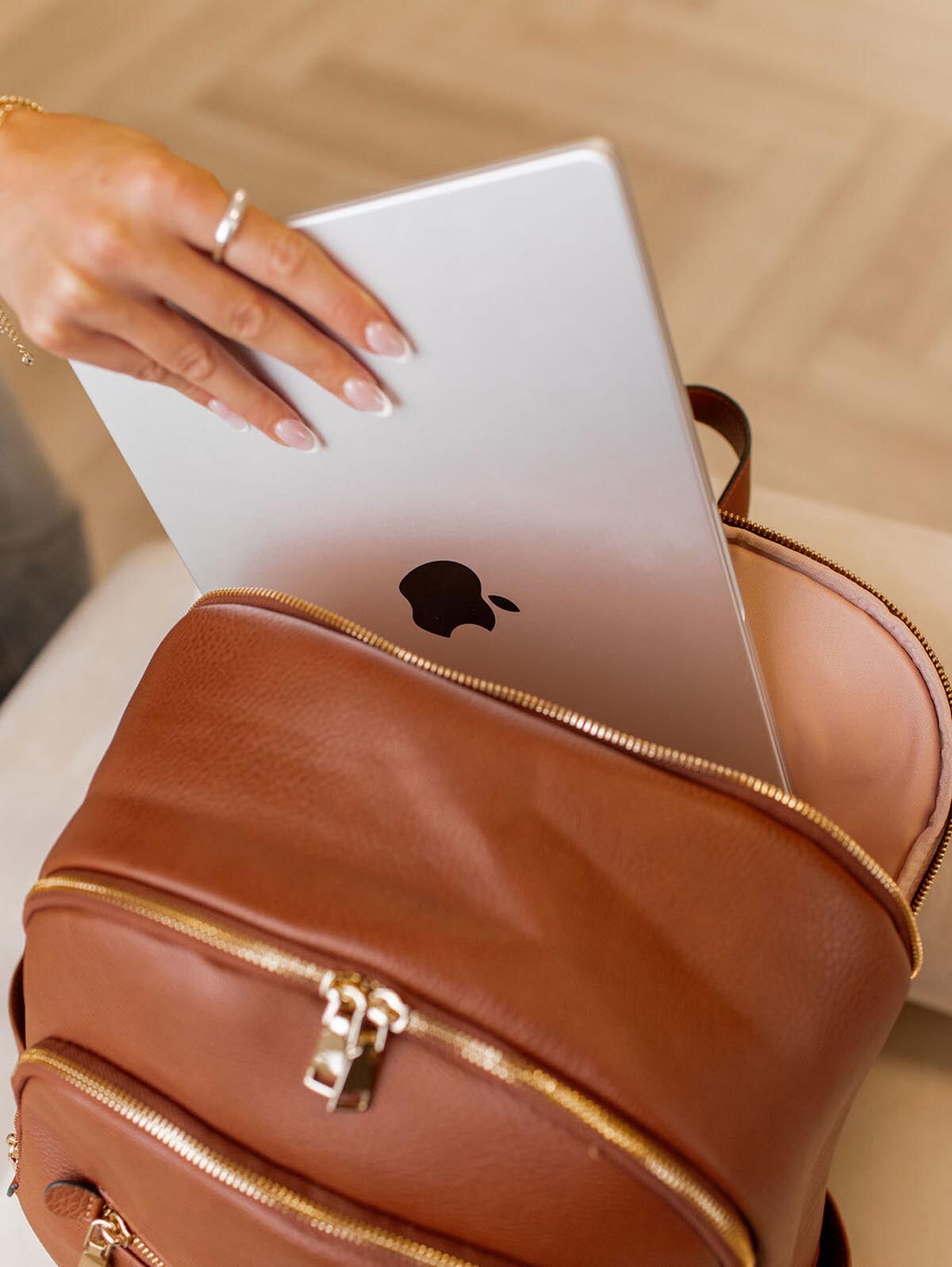 Person putting a silver laptop with an Apple logo into a brown leather backpack.