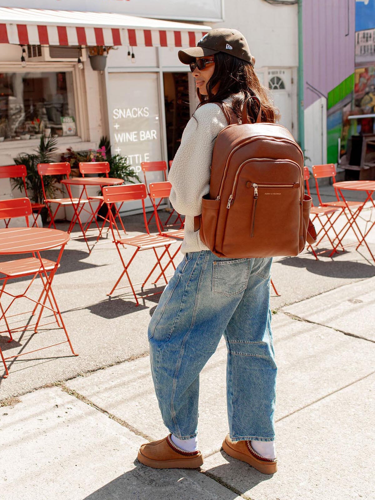 Person with a brown vegan leather backpack walking on a sidewalk with red chairs and tables in the background.